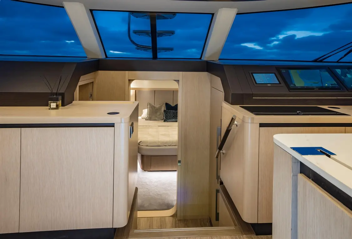 Modern yacht interior featuring a spacious galley, a cozy bedroom through the doorway, and blue skies visible through the window.